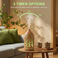 Light wood forest diffuser displaying 3 timer options (1H, 2H, ON) with auto shut-off feature, emitting green mist in a bright living room with green pillows.