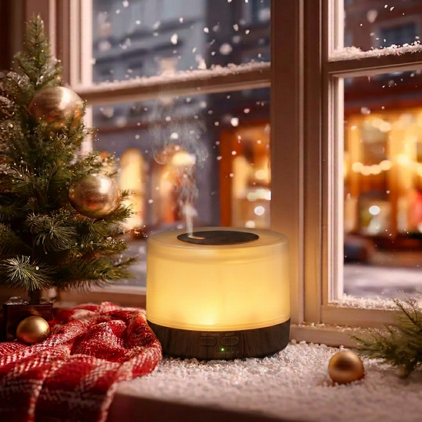Round dark wood ultrasonic aromatherapy diffuser glowing with warm light and mist on a snowy Christmas windowsill, decorated with a small Christmas tree, red plaid blanket, and gold ornaments, creating a cozy winter holiday home ambience and ideal Christmas gift.