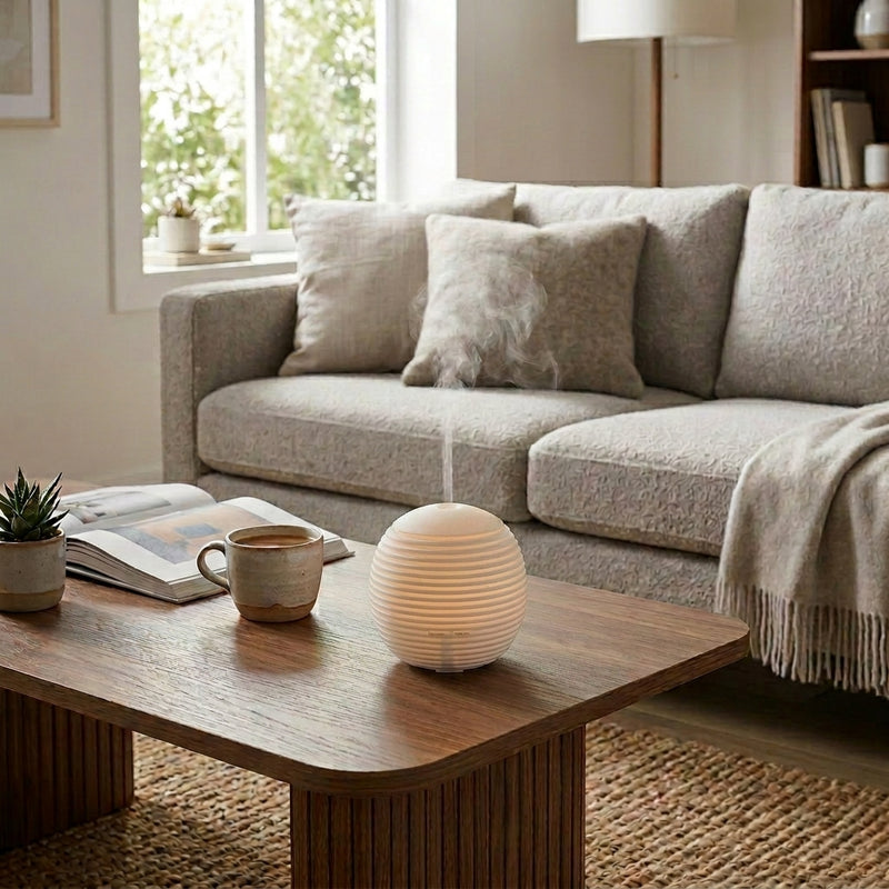 Mini diffuser on a wooden coffee table in a sunlit living room, surrounded by a cup of coffee, an open magazine, and a succulent plant, with a comfortable sofa in the background.