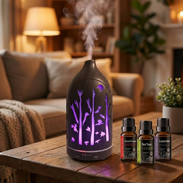 Dark wood forest cutout diffuser emitting purple LED light and mist on a wooden table in a cozy living room, next to a 3-piece essential oil set (Sweet Orange, Tea Tree, Lavender).