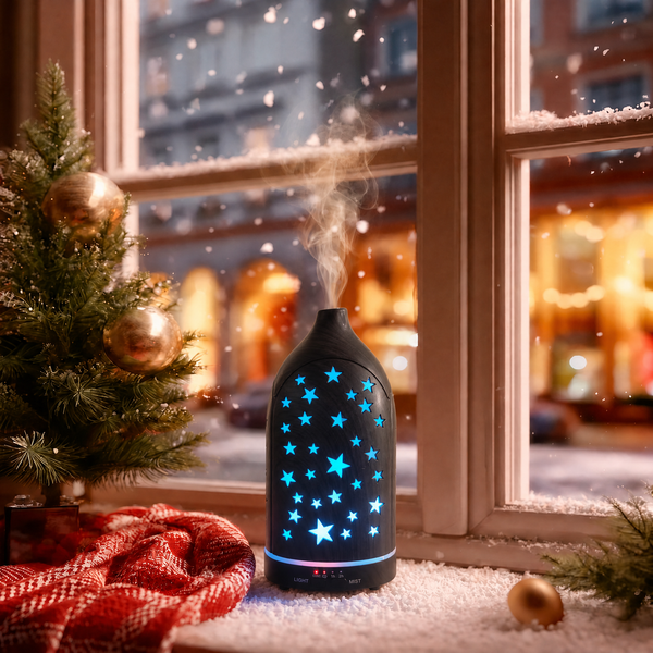 Glowing star cutouts dark wood ultrasonic aromatherapy diffuser with blue LED stars and mist on a snowy Christmas windowsill, decorated with a small Christmas tree, red plaid blanket, and gold ornaments, creating a cozy winter holiday ambience and ideal Christmas gift.