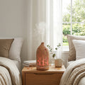 Light wood grain forest diffuser glowing with warm red/orange light on a nightstand between two beds, creating a relaxing bedroom atmosphere with eucalyptus leaves.