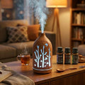 Light wood grain forest cutout diffuser emitting mist and white light on a wooden table with a cup of tea and a 3-pack essential oil set (Lavender, Sweet Orange, Tea Tree).