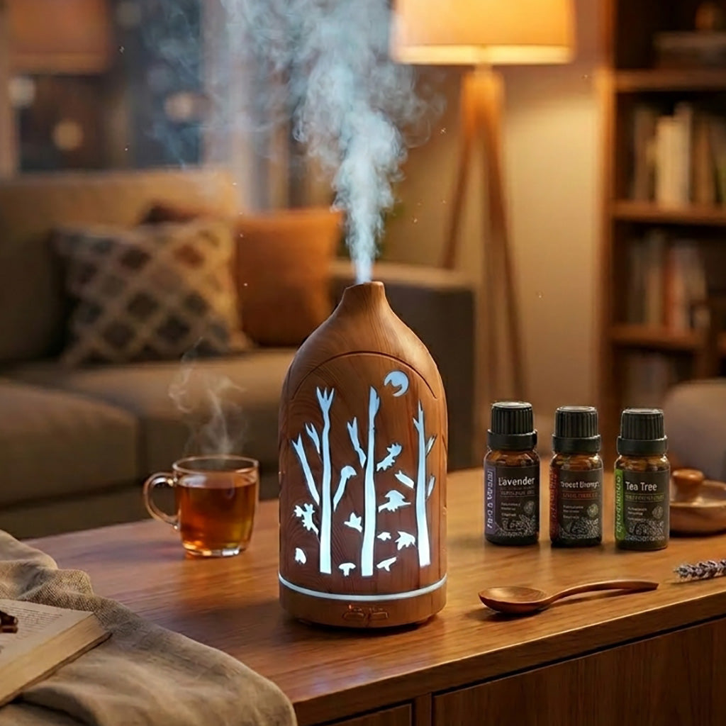 Light wood grain forest cutout diffuser emitting mist and white light on a wooden table with a cup of tea and a 3-pack essential oil set (Lavender, Sweet Orange, Tea Tree).