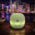 White mini lantern aroma diffuser glowing with green ambient light on a wooden table with a blurred city skyline at night in the background.