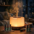 Pniaw dark wood grain round diffuser glowing with warm yellow light at night, surrounded by a 6-bottle essential oil set (Lavender, Peppermint, Lemongrass, etc.) and a wicker basket.