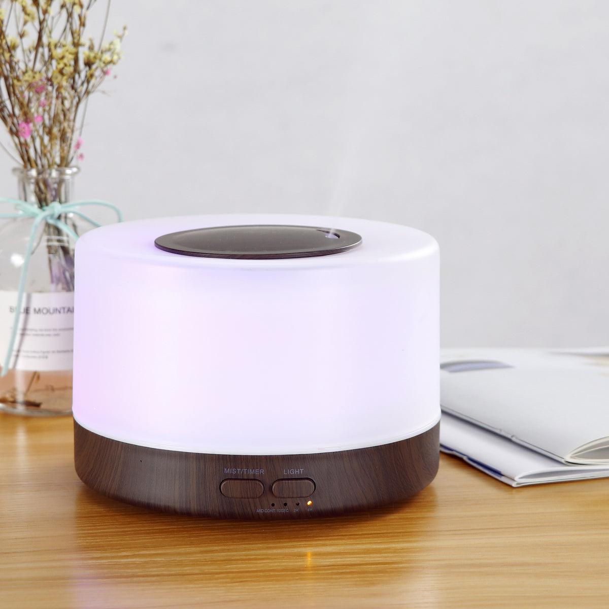Close-up of Pniaw round aroma diffuser with dark wood base and purple LED light, placed on a desk with dried flowers and an open book.